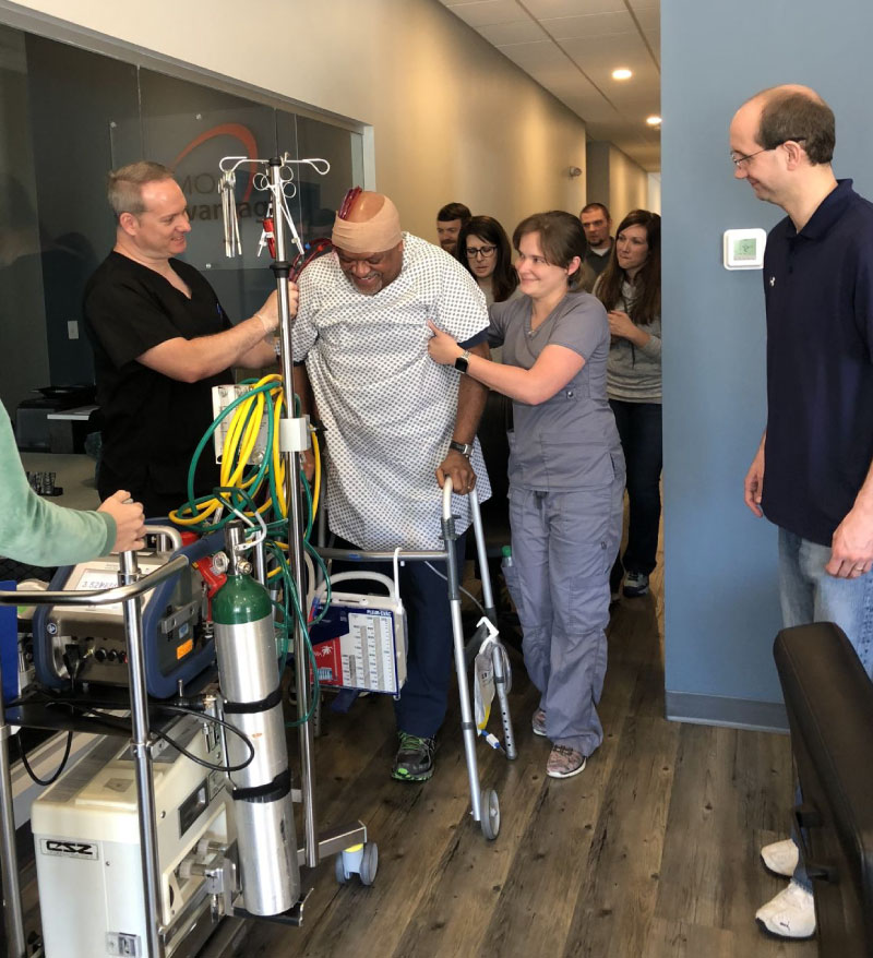 Hospital patient using a walker, attached to tubes and lines, being assisted by hospital personnel
